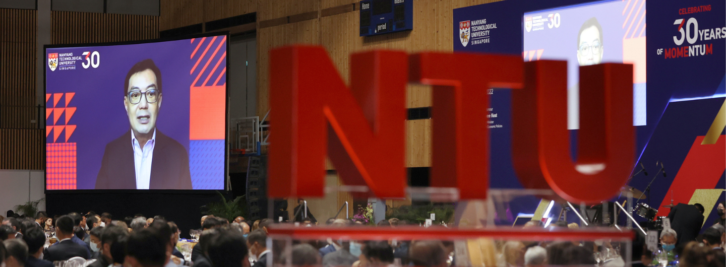 Indoor event with large screen showing speaker, red 'NTU' letters in foreground, audience visible.